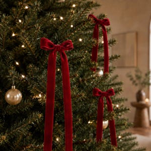 May include: Three deep red velvet ribbon bows attached to a green pine Christmas tree. Each bow features two long, trailing tails hanging vertically. A single smooth, round gold ornament hangs from a branch on the left side of the tree.