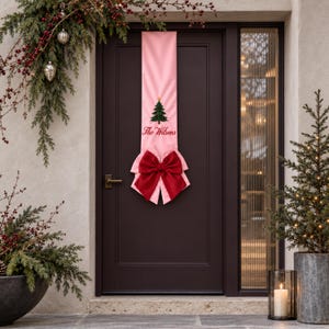 Peut inclure: Une bannière de porte verticale en tissu rose clair ornée d'un sapin de Noël vert avec une étoile dorée au sommet et le texte rouge The Wilsons. La bannière est agrémentée d'un grand nœud en tissu rouge foncé au centre inférieur.