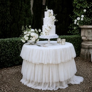 May include: A four-tier white wedding cake with textured frosting and white floral accents sits on a white cake stand. The cake is placed on a round table covered with a layered, ruffled white tulle tablecloth, accompanied by a white floral centerpiece.
