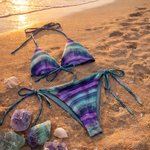 May include: A two-piece bikini set with a striped pattern in shades of purple, teal, and blue, lying on a sandy beach. The bikini top and bottom are tied with matching strings. Several colorful gemstones and a seashell are scattered around the swimwear.