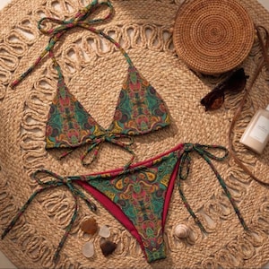 Peut inclure: Un ensemble de bikini à imprimé cachemire coloré, composé d'un haut triangle et d'un bas à nouer. Le bikini est dans les tons de vert, rose et jaune. L'ensemble est présenté sur un tapis tissé, avec des lunettes de soleil, un sac en paille et de la crème solaire.