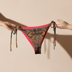 May include: A red bikini bottom with a colorful paisley pattern. The bikini bottom has tie sides and is held up by two hands. The paisley pattern includes shades of green, yellow, and blue.