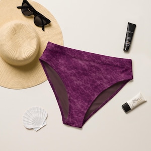 May include: A high-waisted bikini bottom in a deep purple acid-wash fabric, accompanied by a wide-brimmed straw hat, black sunglasses, a tube of sunscreen, and a white seashell. The items are arranged together on a neutral-toned surface.