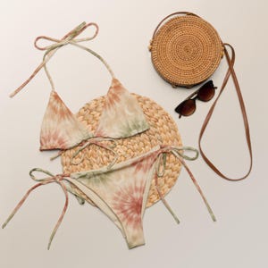 May include: A tie-dye bikini set in shades of green, peach, and cream, with a crescent moon design. The set is displayed with a woven straw mat, a round straw handbag, and brown sunglasses. The bikini top and bottom have tie closures.