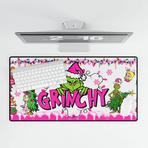Op de afbeelding: Een rechthoekige muismat met een Grinch-thema. Het woord "GRINCHY" is roze en het ontwerp bevat de Grinch met een kerstmuts, kerstverlichting en andere feestelijke elementen. Een wit toetsenbord en muis zijn ook aanwezig.