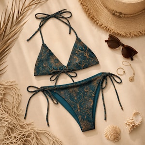 Op de afbeelding: Een teal en goudkleurige bikini set met hemelse thema. De driehoekige top en de broek met strikjes hebben een patroon van manen, sterren en zonnen. Een strohoed, zonnebril, sieraden en schelpen completeren de strand esthetiek.