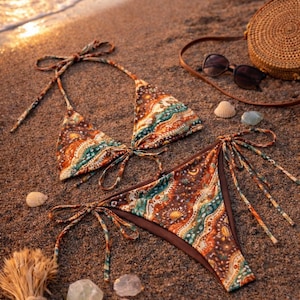 May include: A patterned bikini set with brown, teal, and orange hues, tied with string. The swimwear is displayed on a sandy beach, alongside a woven bag, sunglasses, seashells, and decorative stones.
