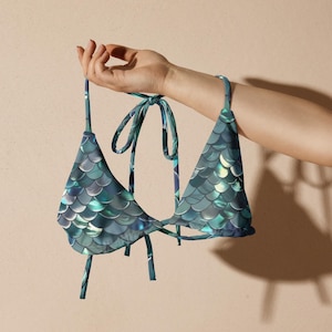 May include: A triangle bikini top featuring a teal and blue iridescent fish scale pattern. The garment includes thin fabric ties at the neck and back, with a classic triangular silhouette and a smooth, reflective finish on the printed fabric.