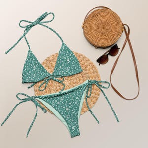 May include: A teal bikini set with a white floral pattern, featuring a triangle top and tie-side bottoms. The set is displayed with a woven straw mat, a round straw bag, and brown sunglasses.