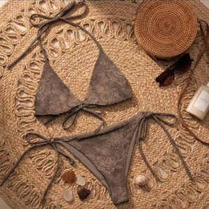 Op de afbeelding: Een tweedelige bikini in een gedempt bruin bloemenpatroon. De top is een haltermodel en de broek is met strikjes. De set wordt getoond op een geweven jute kleed, met een strooien tas, zonnebril en zonnebrandcrème.