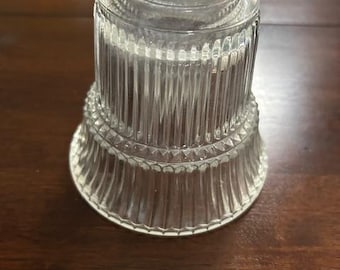 May include: A clear glass light fixture shade with vertical ribbed detailing. The shade has a flared base and a cylindrical top, designed to fit over a light bulb. The glass is transparent, allowing light to pass through. This vintage-style shade is suitable for home lighting.