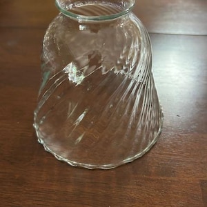 May include: Clear glass lampshade with a spiral design. The lampshade has a flared shape with a scalloped edge. The glass is transparent, allowing light to pass through. The lampshade is designed to diffuse light.