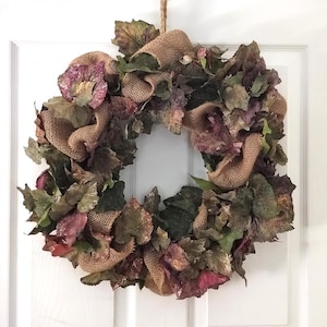 May include: A circular wreath constructed from artificial leaves in shades of deep purple, dark green, and brown, interspersed with loops of tan burlap ribbon. The wreath is suspended by a piece of twine and features a textured, layered appearance.