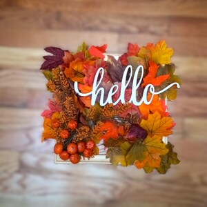 May include: A square autumn-themed decoration with a wooden frame. The frame is filled with colorful fall leaves in shades of orange, red, and yellow. The word "hello" is written in white script across the leaves. Small orange pumpkins are also included.