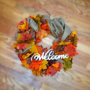 May include: A fall-themed wreath with colorful autumn leaves in shades of red, orange, and yellow. The wreath has a burlap bow and a white "Welcome" sign, perfect for seasonal home decor.