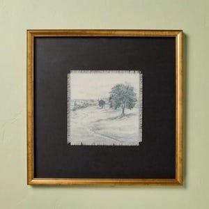 May include: A square framed artwork featuring a pencil sketch of a landscape with a large tree in the foreground and a winding path. The sketch is on a piece of fabric with frayed edges, mounted on a black background inside a gold-toned frame.