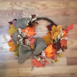 May include: A fall-themed wreath featuring colorful artificial leaves in shades of orange, yellow, and red. The wreath includes a burlap bow and small white floral accents, perfect for autumn decor.
