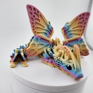 May include: A colorful 3D printed dragon figurine with butterfly wings. The dragon is a light yellow color with a rainbow iridescent finish. The wings are a gradient of pink, yellow, and blue.