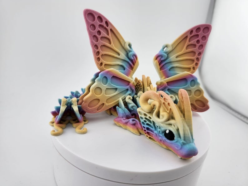 May include: A colorful 3D printed dragon figurine with butterfly wings. The dragon is a light yellow color with a rainbow iridescent finish. The wings are a gradient of pink, yellow, and blue.