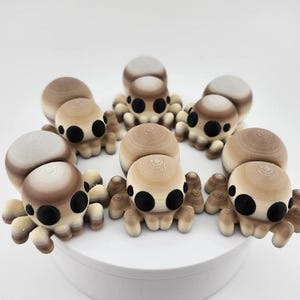 Peut inclure: Six petites figurines en forme d'araignée, de couleur marron et blanche, avec des yeux noirs. Les figurines sont disposées en cercle sur une surface blanche.
