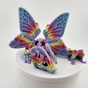 May include: A small, colorful, 3D printed dragon figurine with butterfly wings. The dragon is a light pink, blue, and yellow color with intricate details. The wings are a light purple and blue color with a rainbow gradient.