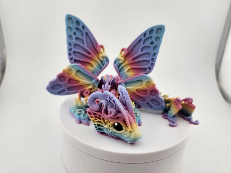 May include: A small, colorful, 3D printed dragon figurine with butterfly wings. The dragon is a light pink, blue, and yellow color with intricate details. The wings are a light purple and blue color with a rainbow gradient.