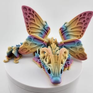 May include: A colorful 3D printed dragon figurine with butterfly wings. The dragon is a light yellow color with blue and pink accents. The wings are a gradient of pink, yellow, and blue.