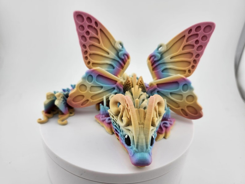 May include: A colorful 3D printed dragon figurine with butterfly wings. The dragon is a light yellow color with blue and pink accents. The wings are a gradient of pink, yellow, and blue.