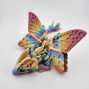 May include: A 3D printed dragon figurine with iridescent wings, resembling a butterfly. The dragon is made of a light beige plastic with a rainbow color gradient. The wings are translucent and have a delicate, intricate design.