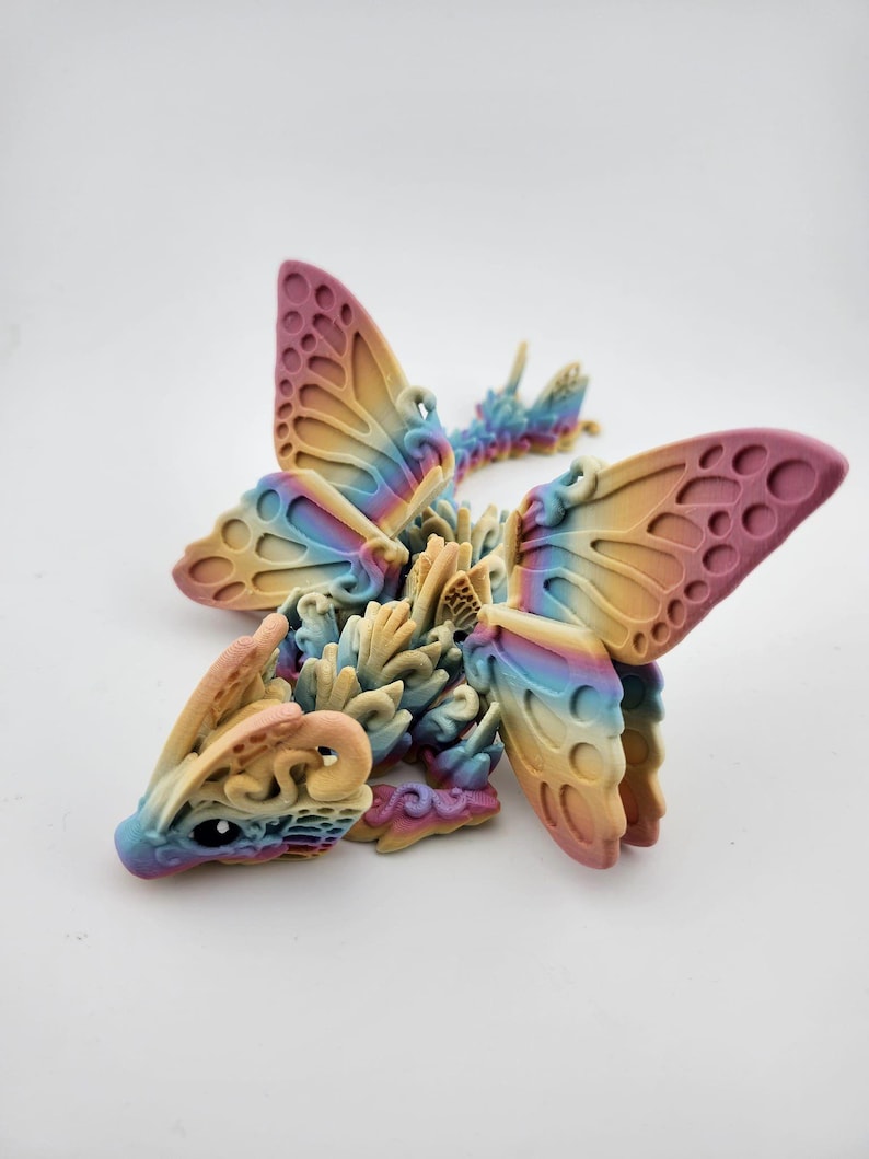 May include: A 3D printed dragon figurine with iridescent wings, resembling a butterfly. The dragon is made of a light beige plastic with a rainbow color gradient. The wings are translucent and have a delicate, intricate design.