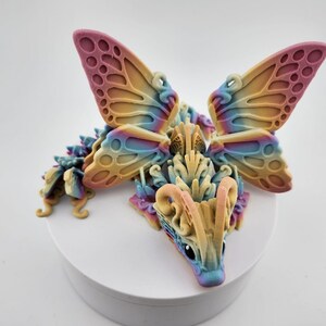 May include: A colorful 3D printed dragon figurine with butterfly wings. The dragon is a light yellow color with blue and pink accents. The wings are a gradient of pink, blue, and yellow.