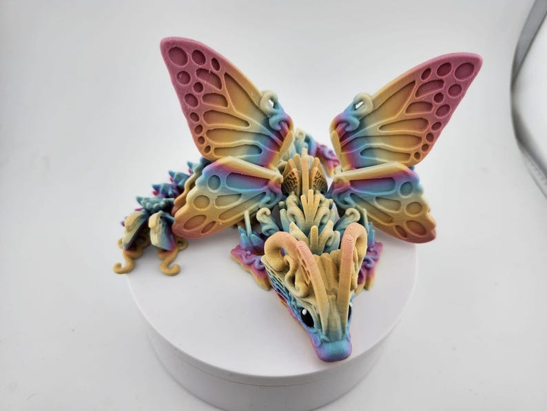 May include: A colorful 3D printed dragon figurine with butterfly wings. The dragon is a light yellow color with blue and pink accents. The wings are a gradient of pink, blue, and yellow.