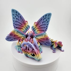 May include: A rainbow-colored 3D printed dragon figurine with butterfly wings. The dragon is sitting on a white circular base.