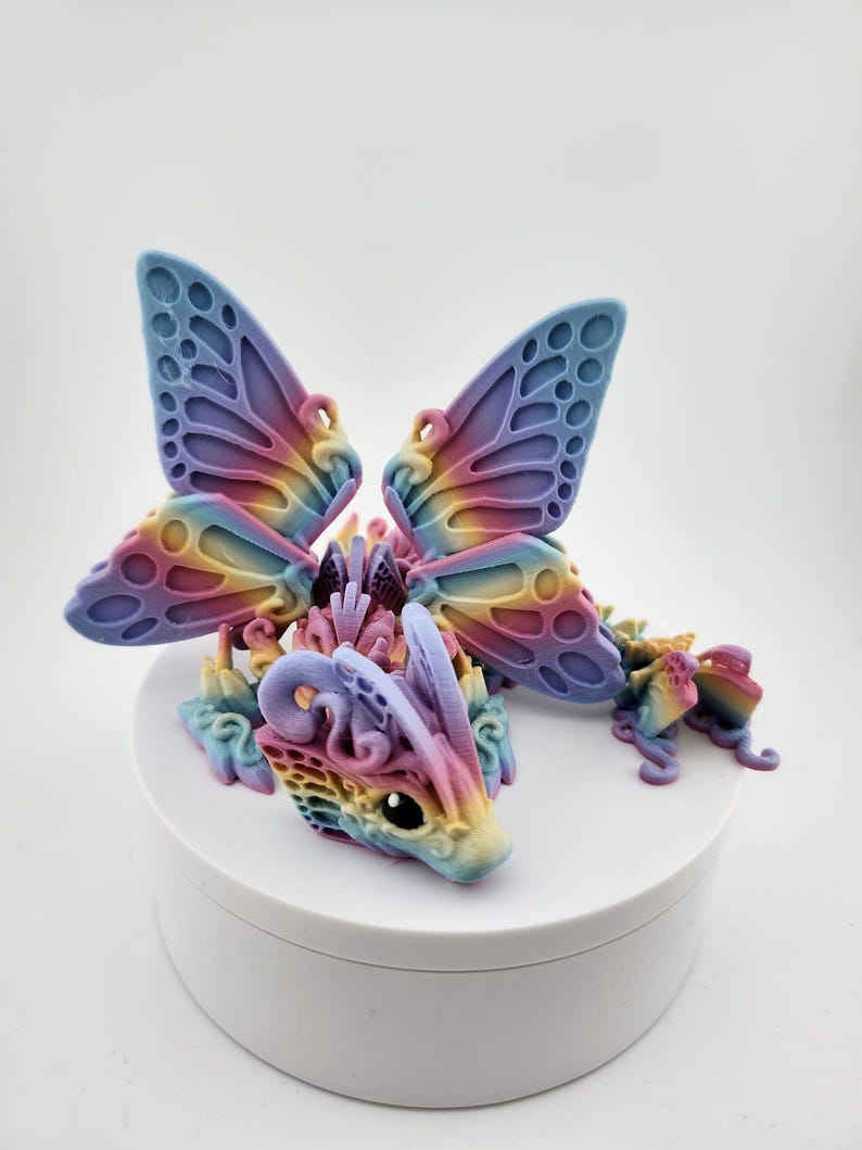 May include: A rainbow-colored 3D printed dragon figurine with butterfly wings. The dragon is sitting on a white circular base.