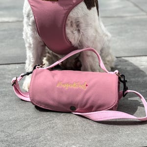 Puede incluir: Un bolso rosa para perros con un arnés a juego. El bolso tiene forma cilíndrica con una correa para el hombro y el texto "Huge & Ted" bordado en dorado. El arnés es visible en el fondo.