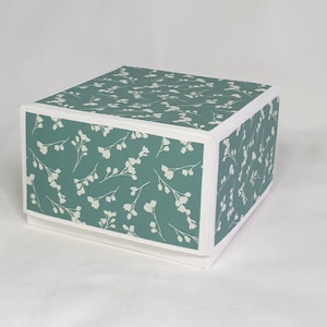 Op de afbeelding: Een vierkante teal gekleurde geschenkdoos met een wit bloemenpatroon en een witte rand.