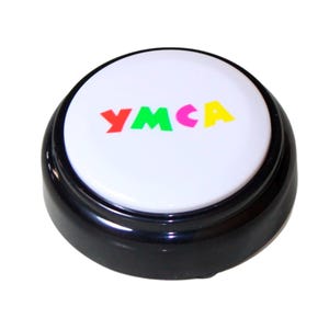 Op de afbeelding: Een ronde, zwarte knop met een witte bovenkant. De bovenkant toont het woord "YMCA" in rode, groene, gele en blauwe blokletters. Deze knop is waarschijnlijk een novelty item of een speelgoed.