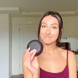 Peut inclure: Un compact circulaire brun foncé tenu dans la main d'une personne. Le compact semble être fait d'un matériau mat. La personne porte un débardeur bordeaux. L'arrière-plan comprend un mur blanc, une porte et une commode.