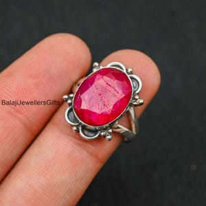 May include: A silver ring featuring a large, oval ruby gemstone. The ring has a delicate, ornate design with small silver beads and swirls.