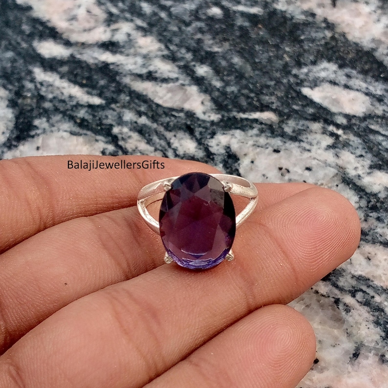 May include: A silver ring featuring a large, oval-cut purple gemstone. The ring has a simple band with a split design on either side of the gemstone. The text "BalajiJewellersGifts" is visible.