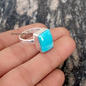 Może przedstawiać: Srebrny pierścionek z kwadratowym, jasnoniebieskim kamieniem szlachetnym. Pierścionek ma gładką, prostą obrączkę, a kamień jest osadzony w srebrnej oprawie. Na pierścionku widoczny jest napis "BalajiJewellersGifts".