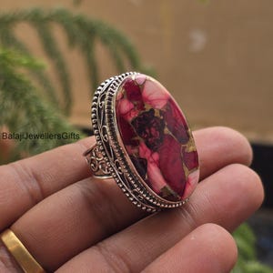 May include: A silver ring with an oval-shaped stone. The stone features a mix of red, pink, and gold colors. The ring has an ornate, detailed design around the stone and band. The ring is held in a hand.