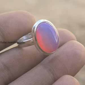 May include: A silver ring with an oval, color-changing stone. The stone transitions from pink to purple. The ring is held in a hand, with the brand name "BalajiJewellersGifts" visible on the ring's band.