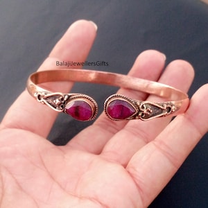 May include: Copper cuff bracelet featuring two teardrop-shaped, deep red gemstones. The open-ended design has ornate detailing near the gemstones. The bracelet is held in a hand, showcasing the jewelry's design and color.