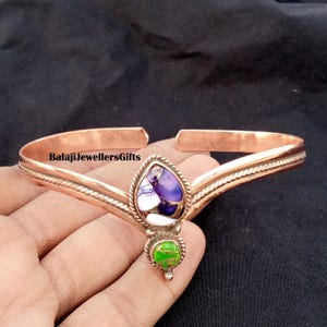 Op de afbeelding: Koperkleurige armband met een centrale druppelvormige steen met paarse, witte en groene accenten. De armband heeft een textuurontwerp en de tekst "BalajiJewellersGifts".