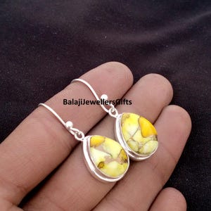 May include: A pair of silver teardrop earrings with a yellow and brown stone inlay. The earrings have a simple hook design and are displayed against a dark background. The text "BalajiJewellersgifts" is visible.