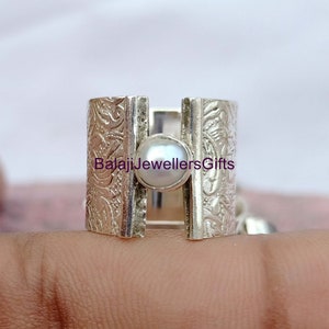 以下が含まれることがあります： 装飾的なバンドに白い真珠がセットされたシルバーリング。バンドは花柄のデザインで、中央に分割されたセクションがあります。