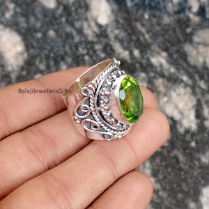 May include: A sterling silver ring with an oval, faceted, light green gemstone. The ring has an ornate design with scrollwork and beaded accents. The text "BalajiJewellersGifts" is visible on the ring.