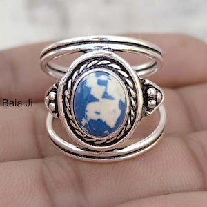 Puede incluir: Anillo de plata con una piedra ovalada azul y blanca. La piedra está engastada en un marco decorativo con un borde trenzado. El anillo tiene un diseño de doble banda y es probablemente una joya.