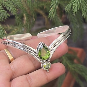 May include: A silver-toned bracelet with a central design featuring a pear-shaped green gemstone and a smaller, round green gemstone below. The bracelet has an open design and is held by a hand. The text "BalajiJewellersGifts" is visible.